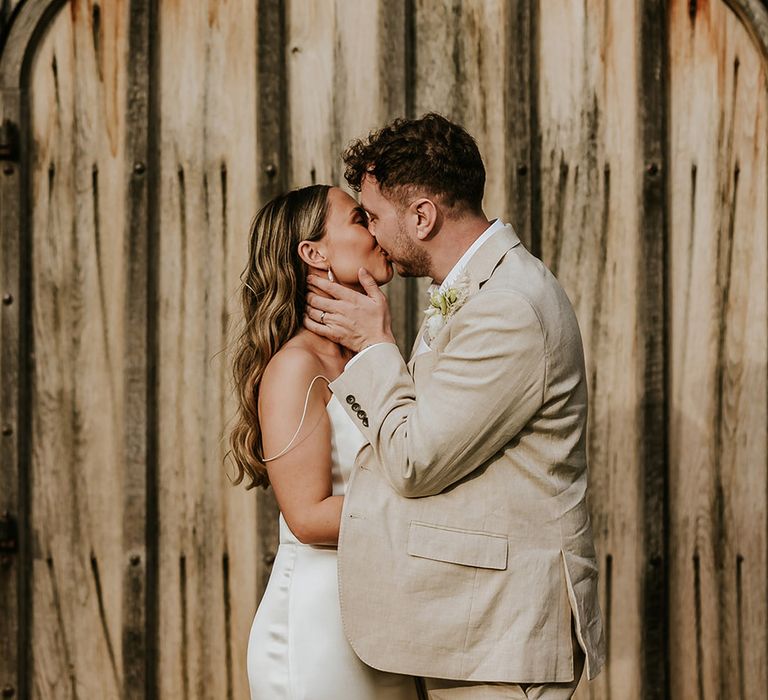 Groom in beige wedding suit kissing the bride wearing a satin slip wedding dress 