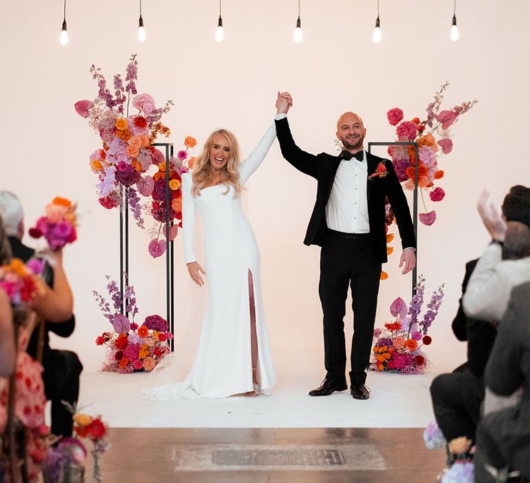 Bride wearing long sleeve Suzanne Neville wedding dress with shoulder pads with groom in black tuxedo facing their guests as a married couple