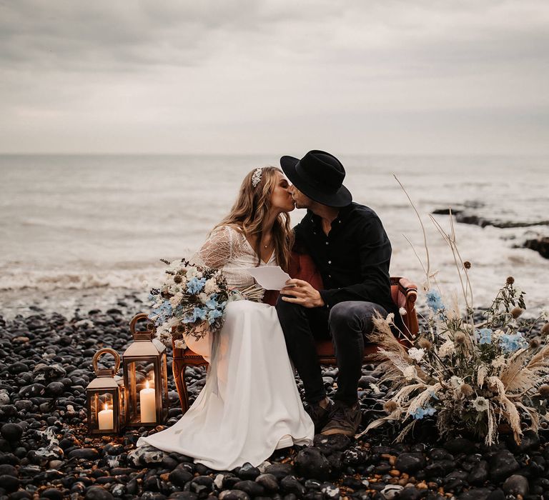 Bride and groom portrait on the beach with blue and white flowers, lanterns and love seat