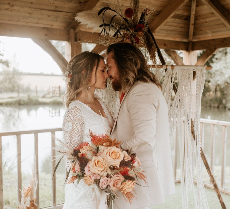 Bride & groom kiss with lake behind under wooden roof