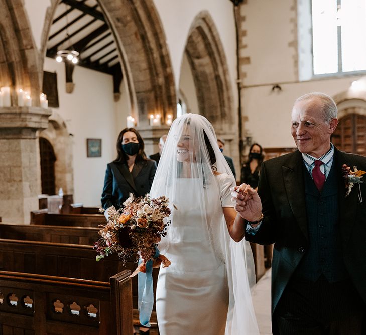 Bride walks down the aisle holding hands with her father. She has a veil over her face and holds her bouquet.