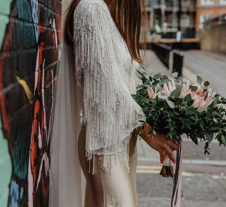 Bride wearing wedding dress with white tassel sleeves holds protea bouquet