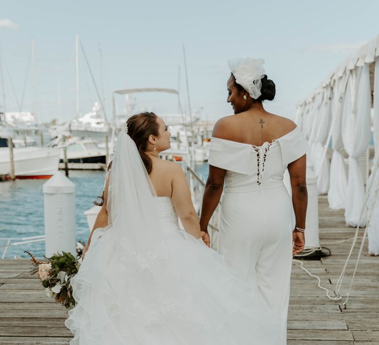 Black bride in Bardot jumpsuit holding her brides hand on the marina dock