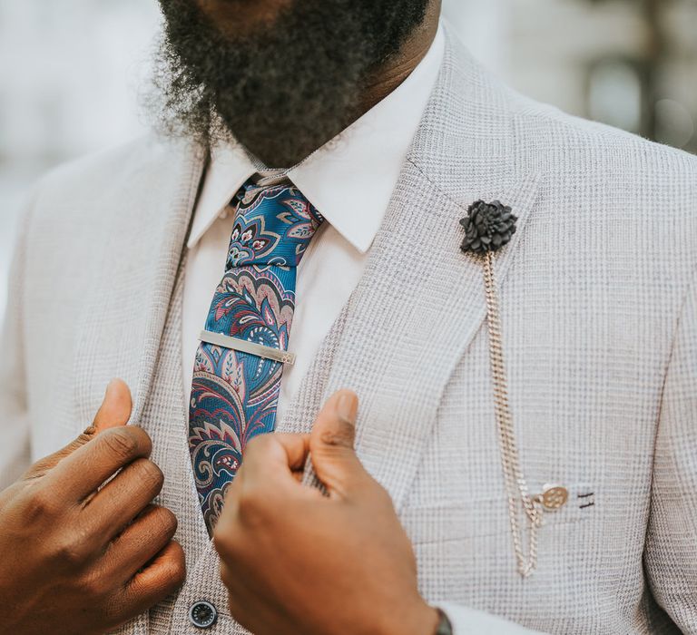 Black groom in light grey checked suit with paisley blue tie, silver tie pin and buttonhole chain 