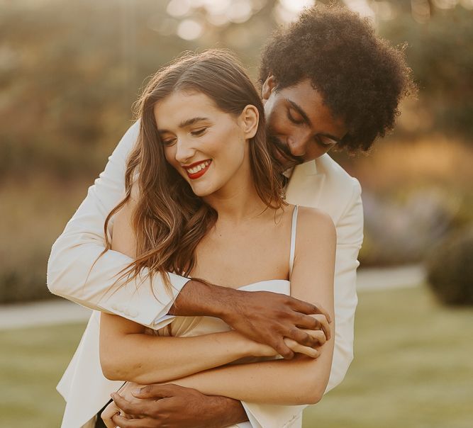 Black groom with afro hair and white tuxedo jacket embracing his bride with long brown hair and red lipstick