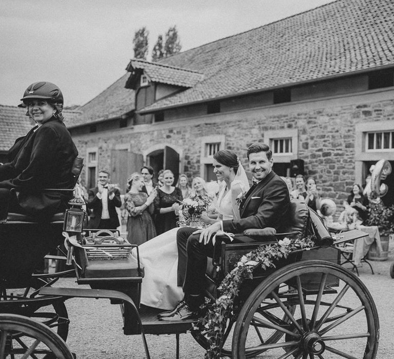 Smiling bride and groom arrive in open horse-drawn carriage to wedding reception at rural German wedding