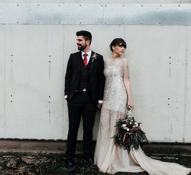 A bride and groom stand side by side, She holds an oversized homemade wedding bouquet