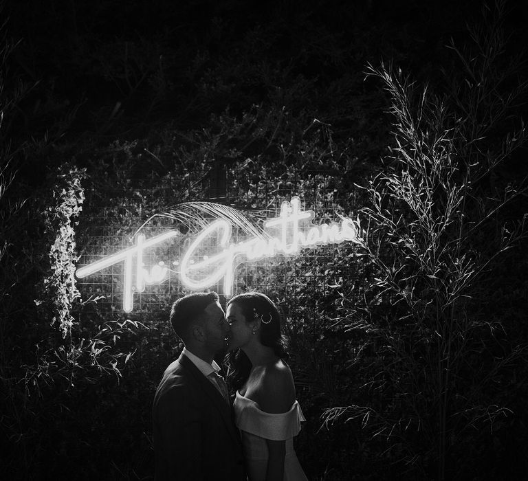 Bride in strapless Vagabond wedding dress kisses groom in grey Hugo Boss suit in front of neon wedding sign 