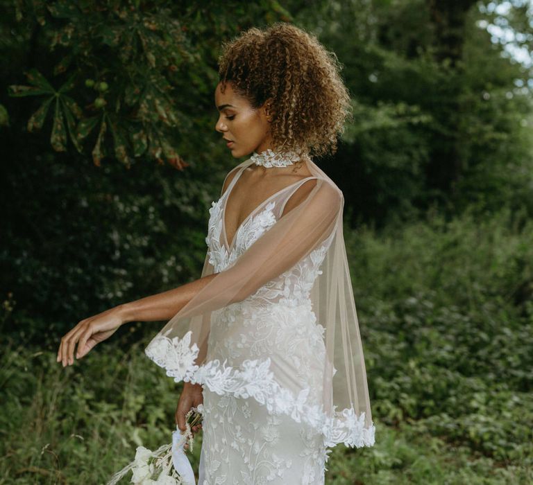 Bride wearing a floral embroidered wedding dress with matching tulle bridal cape in the woods for boho wedding at Berwick Lodge