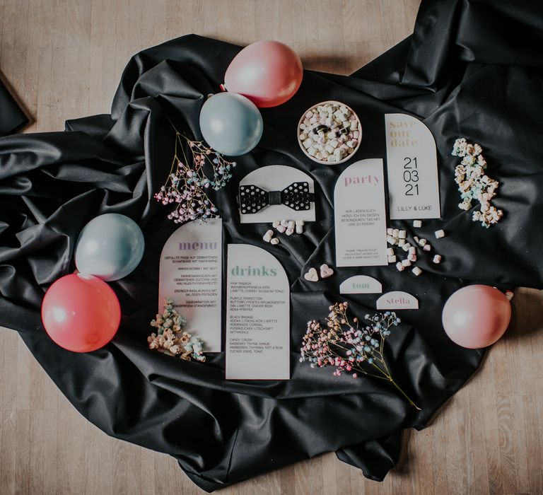 Flat lay of white acrylic wedding stationery suit resting on a black satin tablecloth surrounded by pastel balloons and flowers 