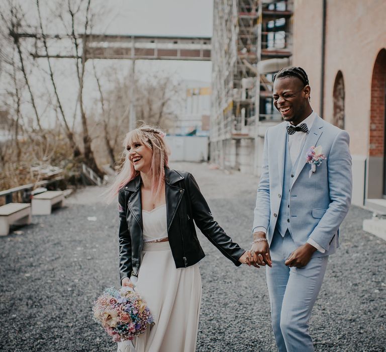 Groom in a pale blue wedding suit laughing with his bride in a leather jacket holding a pastel flower bouquet 