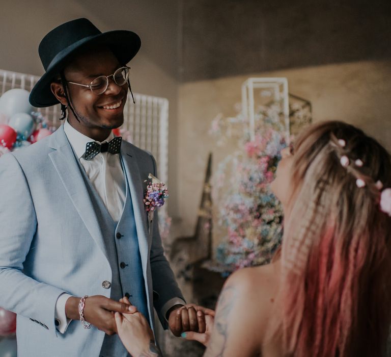 Groom in a pale blue wedding suit, black hat and metal bow tie holding hands with his bride