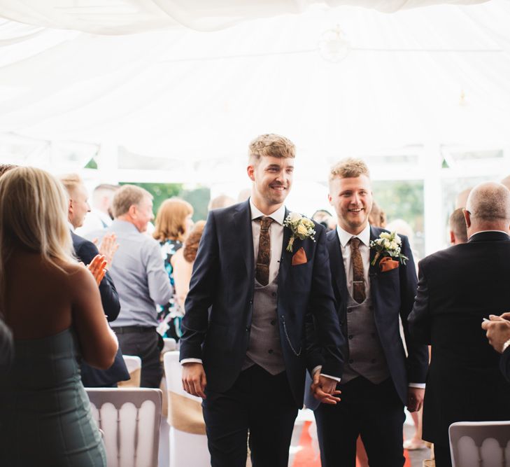 A gay couple leave their wedding ceremony hand in hand wearing matching three-piece suits. Photography by Cat Lane. 