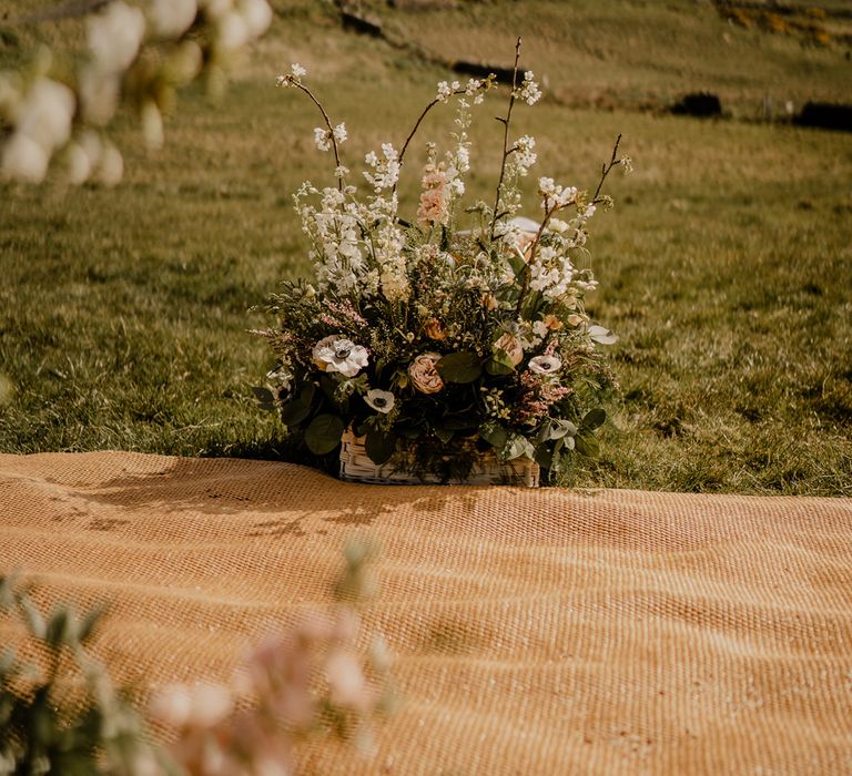 Hessian mat aisle in field with white, pink and green floral arrangement in white woven basket at clifftop ceremony for Dunluce Castle wedding