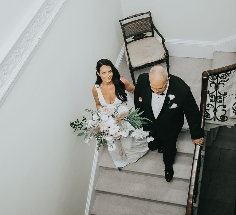 Bride with long dark curled wedding hair wearing a white Pronovias wedding dress with train walks down stairs arm in arm with man in tuxedo as she holds wedding bouquet and looks up