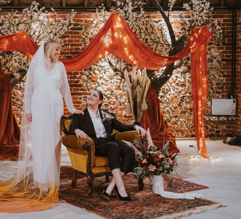 Two brides in front of indoor blossom trees, with vintage rugs and armchair. One bride is wearing a plum coloured suit, the other a long sleeve beaded gown with long bridal cape dip dyed orange