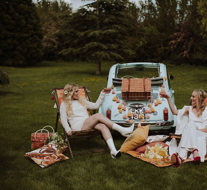 Sixties wedding inspiration with two brides sitting on deck chairs enjoying a picnic by their fiat 500