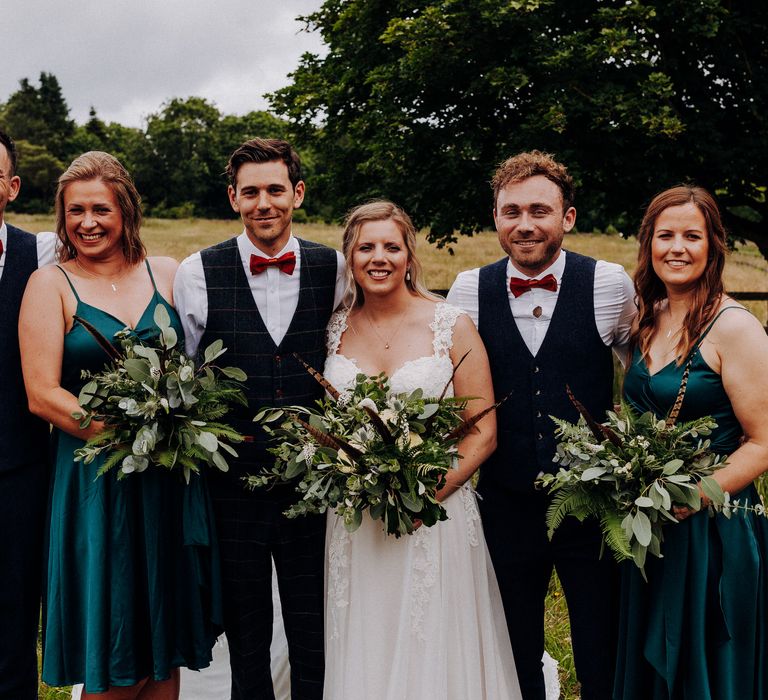 Bride & groom stand with bridesmaids and ushers on their wedding day as bridesmaids wear deep green satin dresses 