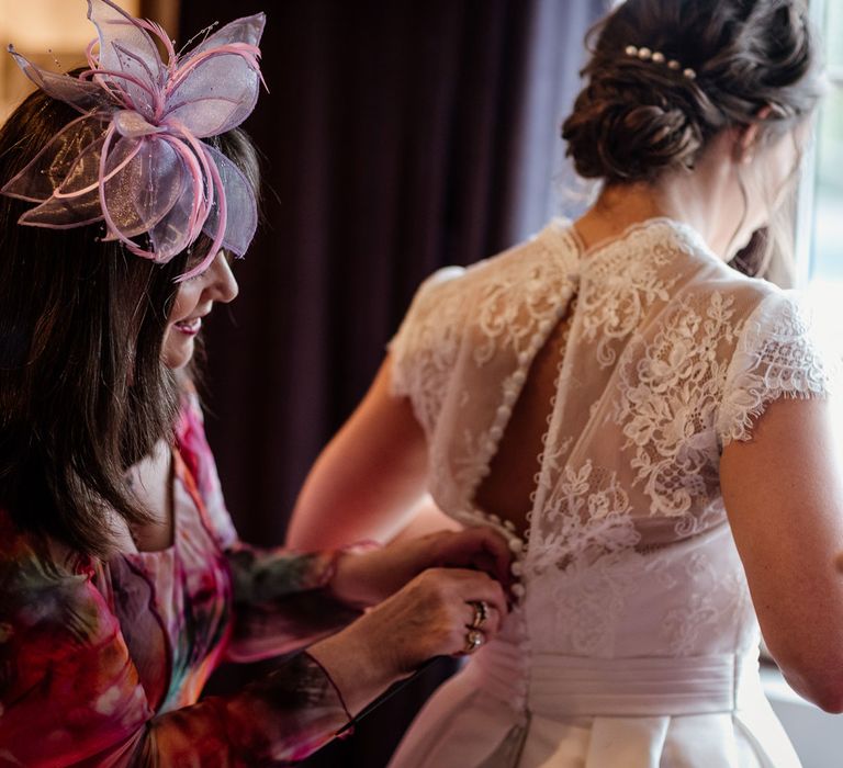 Bride with wedding hair updo wearing lace cap sleeved wedding dress gets her buttons fastened by wedding guest in colourful dress and fascinator at The West Mill Derby