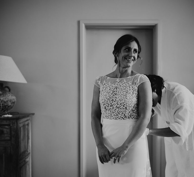 Bride with wedding updo in Rime Arodaky wedding dress with lace train gets the back of her dress done up by guest in dressing gown 