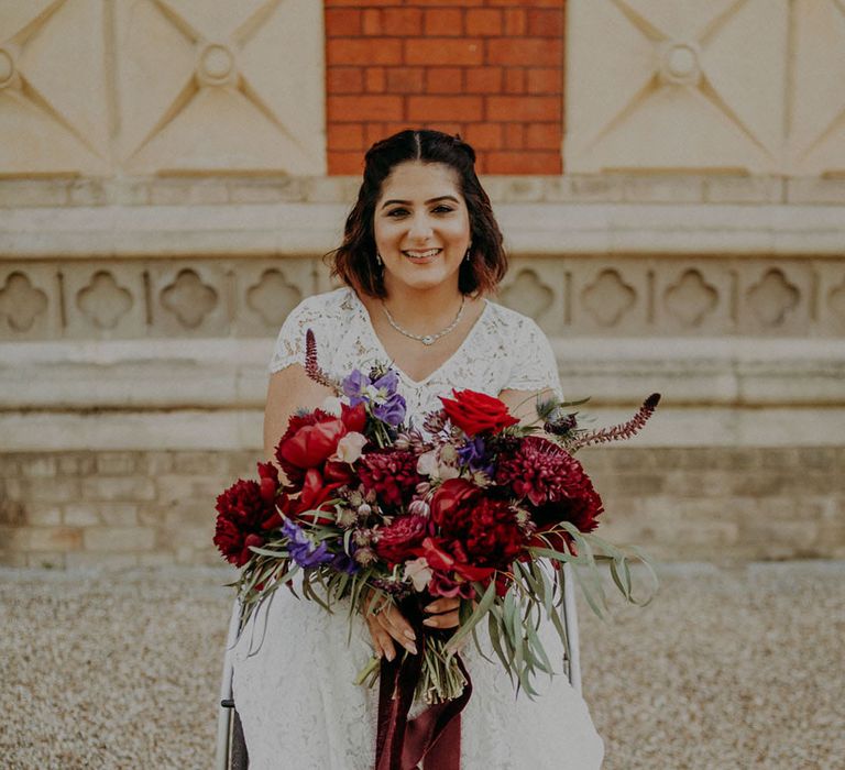 Indian bride in a wheelchair holding her jewel tone red, purple and pink wedding bouquet wearing a lace dress and embellished shoes 