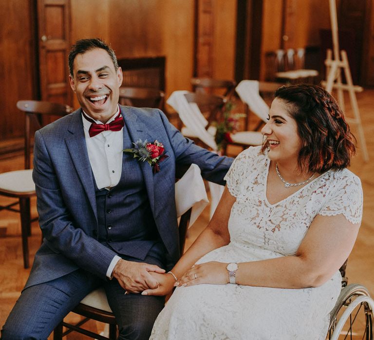 Wedding ceremony at Dulwich College with groom in a blue suit laughing and holding hands with his bride in a wheelchair wearing a lace wedding dress