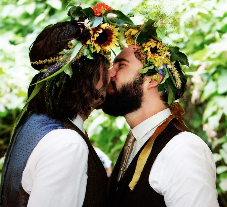 Grooms kiss on their wedding day as they wearing floral gowns and waistcoats for midsummer vibe wedding