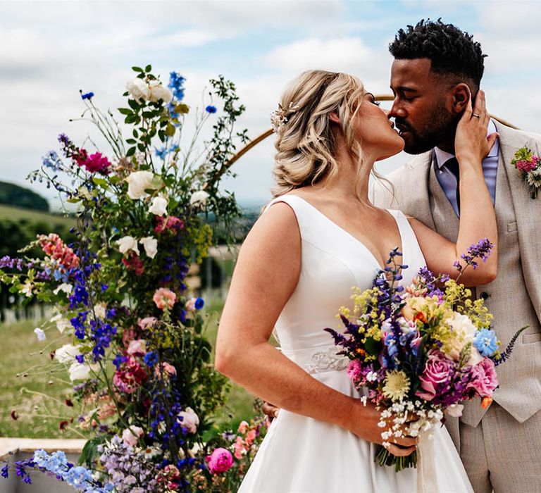 Black groom in a beige three-piece wedding suit kissing his bride in a princess wedding dress as she holds a colourful wildflower bouquet 