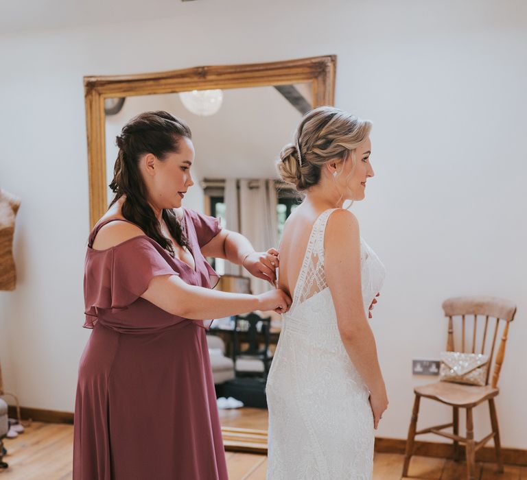 Bride in lace Justin Alexander wedding dress with train and low back gets buttoned up by bridesmaid in dusky pink bridesmaid dress before Tythe Barn wedding with barn wedding flowers