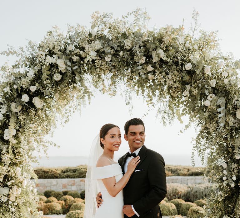 Bride & groom stand beneath floral installation at the end of the aisle on their wedding day | Hannah MacGregor Photo & Film