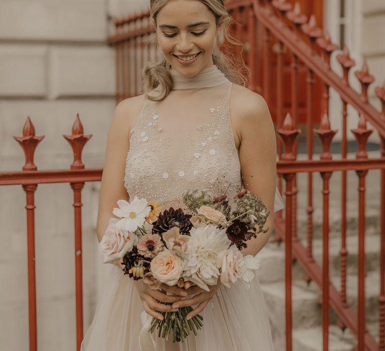 Beautiful bride in a halterneck tulle wedding dress with embellished front holding a autumn wedding bouquet with roses and dahlias 