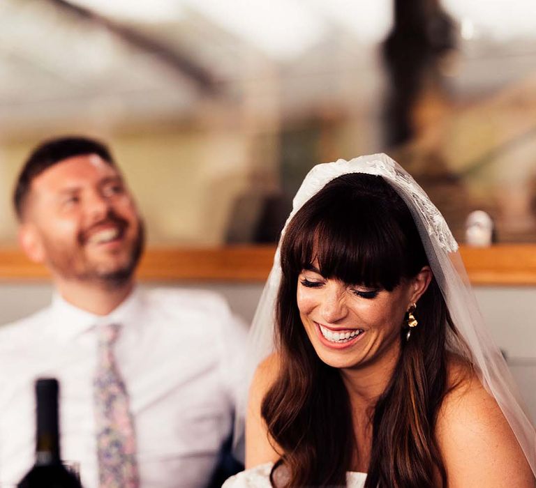 Beautiful bride with a fringe and long wavy hair wearing a veil laughing during the wedding speeches 