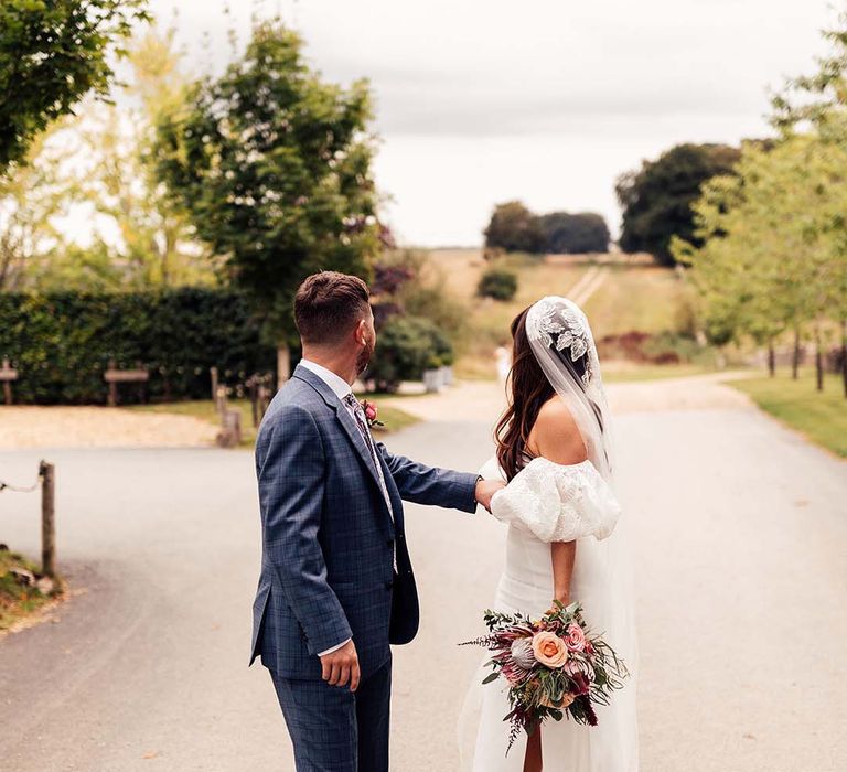 Groom in a blue check suit and bride in a strapless wedding dress with lace detachable sleeve and veil at their Stone Barn Cotswolds wedding 