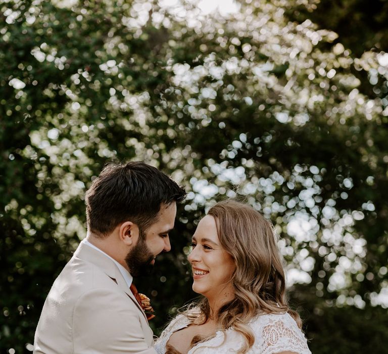 Bride in homemade lace wedding dress holding mixed rose bridal bouquet with curled hair smiles at groom in beige suit with floral buttonhole at DIY garden wedding ceremony