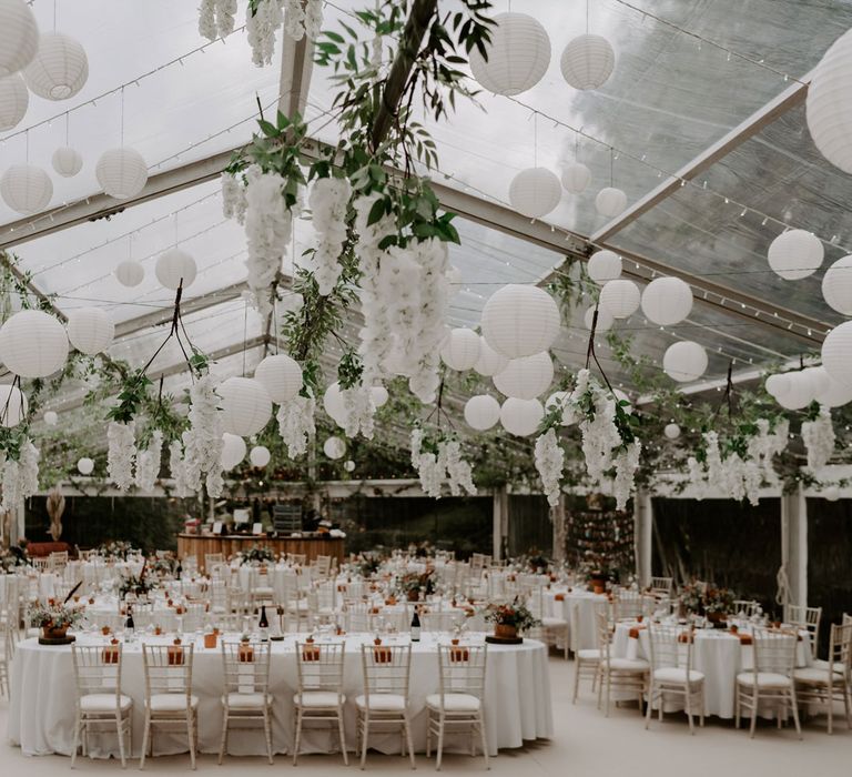 Interior of clear-roofed marquee in garden with white paper lanterns and garlands hanging from the ceiling over white tables and chairs with plant decor for home garden wedding