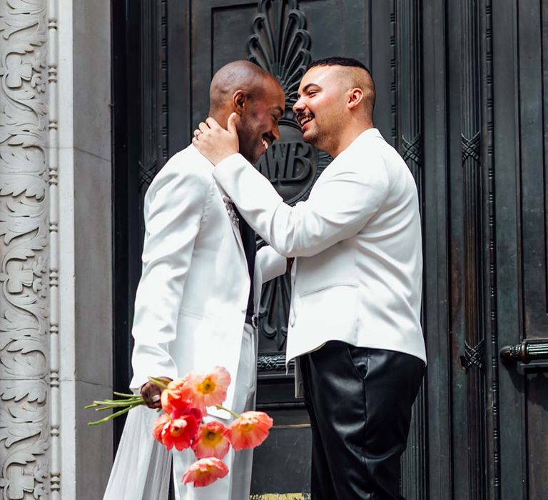 Two grooms at city elopement with groom in leather trousers and white blazer, and groom in a white suit with cape holding a red bouquet 