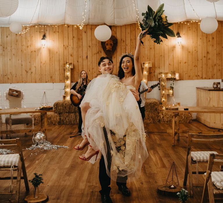 Groom in dark suit holding bride in strapless tulle wedding dress holding up bouquet for rustic wedding reception with fairy lights