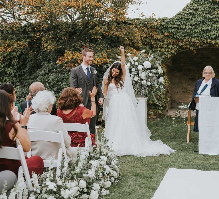 Bride in applique Pronovias wedding dress and veil throws hand in the air as she stands holding hands with groom in grey three piece suit during outdoor wedding ceremony in Buckinghamshire