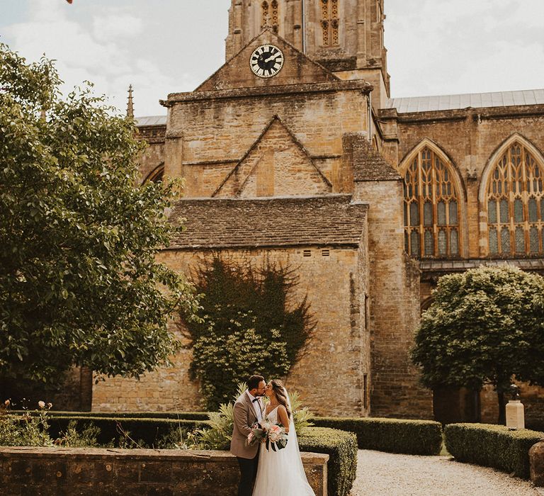 Bride & groom stand and kiss outside church as bride holds tropical inspired bouquet complete with palm leaves and mini pineapples 