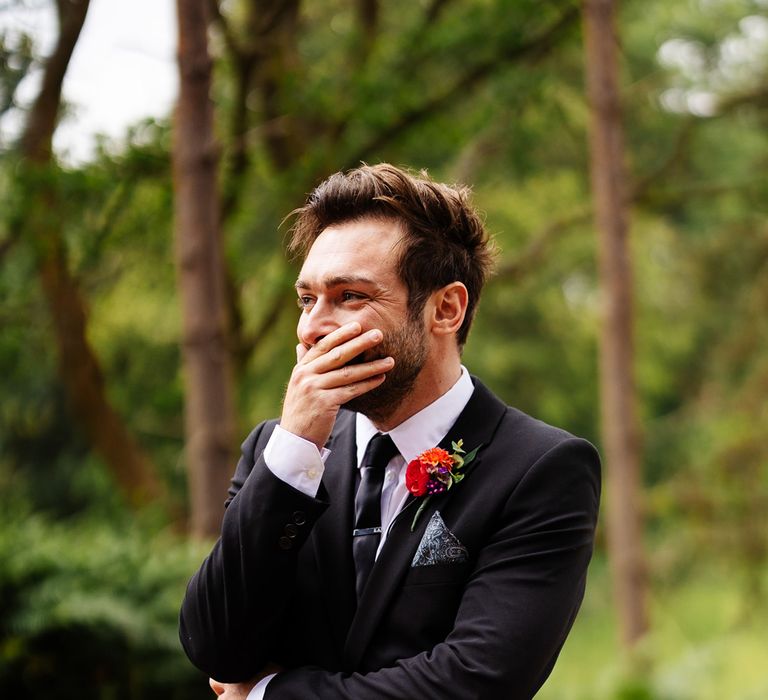 Groom in black suit and colourful floral buttonhole holds hand over his mouth as he smiles at bride walking down the aisle during outdoor woodland wedding ceremony