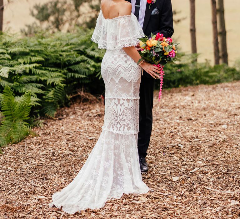 Bride in lace off the shoulder Grace Loves Lace wedding dress and flower crown holds colourful wedding bouquet as she stands smiling with groom in black suit in woodland