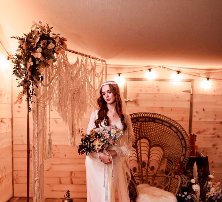 Bride in long lace sleeve wedding dress with Juliet cap veil stands in wooden cabin with rattan chair, string lights and candles