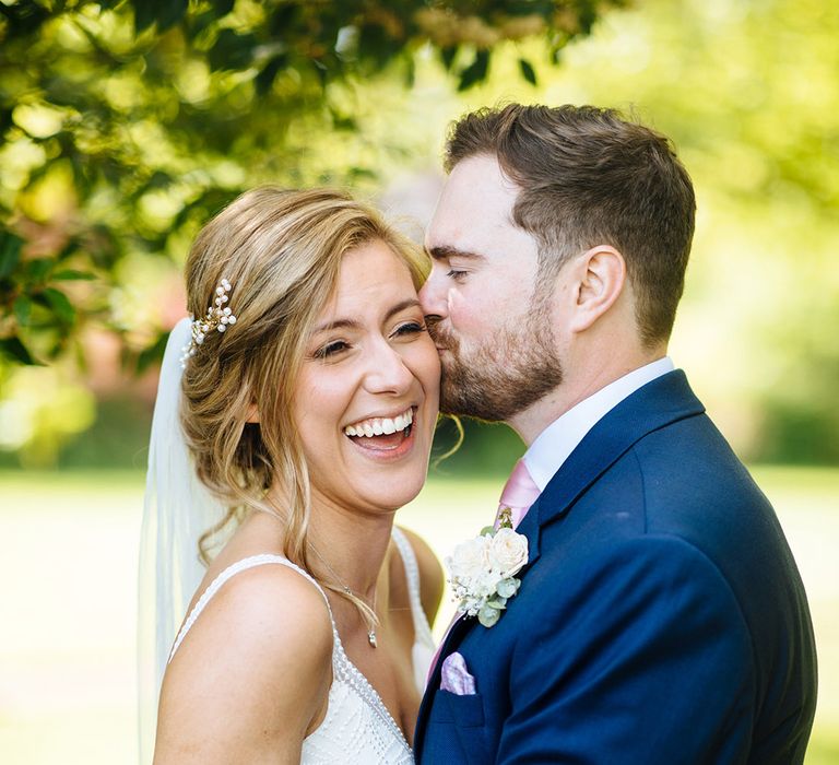 Bearded groom in a navy suit kissing his brides cheek with a pinned up do, cathedral length veil and Made With Love Bridal gown 