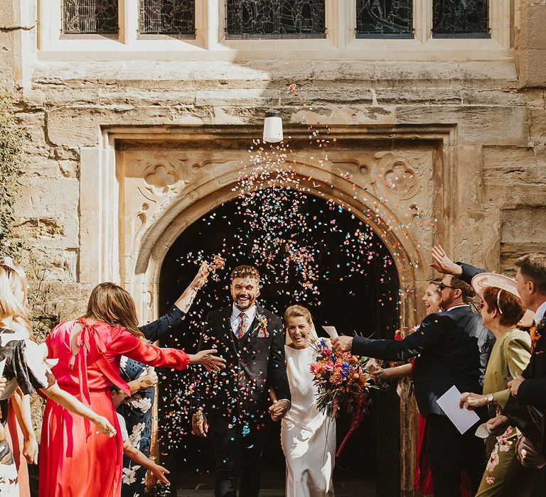 Colourful couch confetti exit with groom in a navy three-piece suit and bride in a Charlie Brear wedding dress