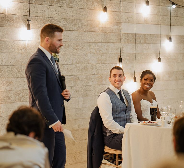 Best man stands to do his speech at wedding reception full of hanging lights to create the perfect setting