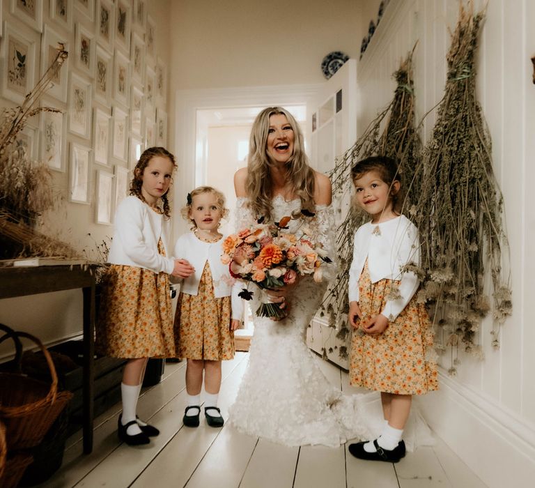 Bride in off the shoulder wedding dress with applique flowers holding rustic bridal bouquet stands with flower girls in sunflower dresses and cardigans in hallway