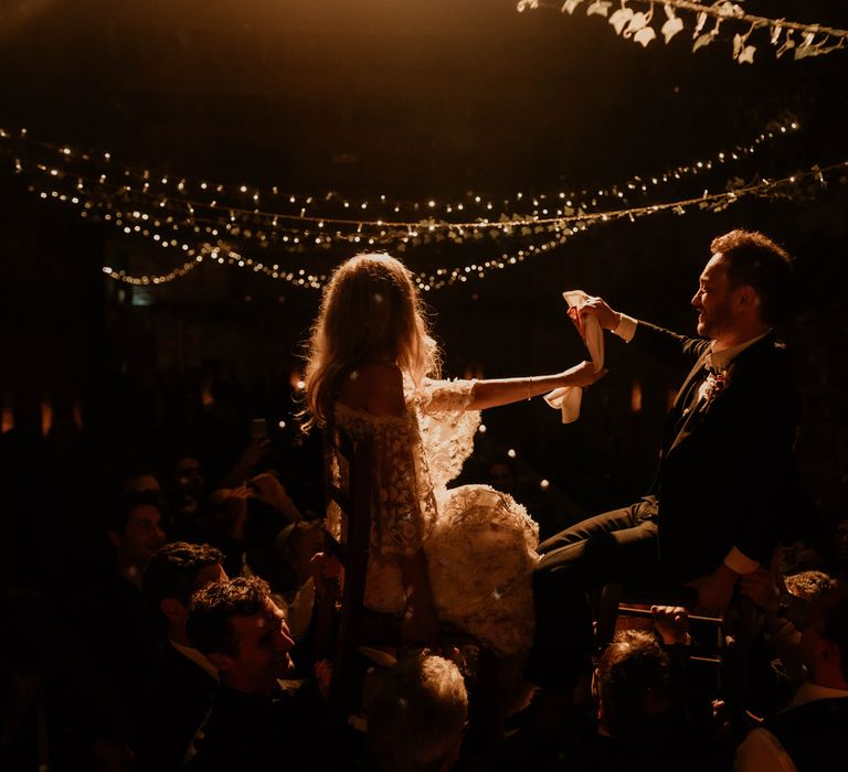 Bride in boho wedding dress and groom in dark suit sit on chairs during horah at evening wedding reception 