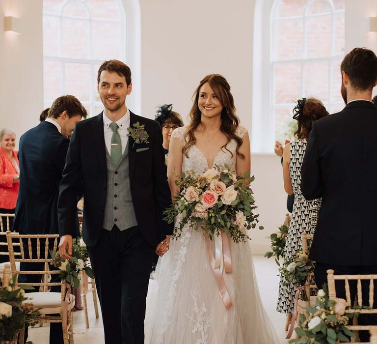 Bride in Madi Lane wedding dress clutches blush bouquet holding grooms hand