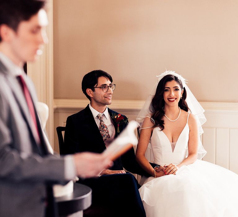 Bride in white wedding dress with plunging neckline, pearl necklace and veil  sits with groom in dark suit and floral Gucci tie whilst wedding guest performs a wedding reading