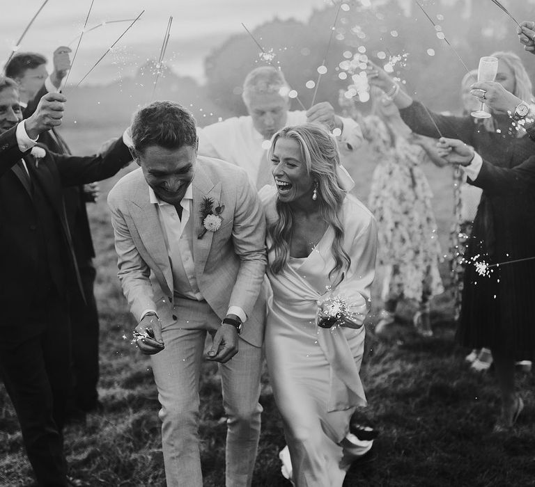 Laughing bride in wrap top and groom in open shirt and suit laugh as they have sparkler exit in field after garden wedding reception